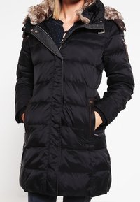 Svart vinterjacka med fuskpälsfodrad huva, dragkedjestängning, två sidofickor och strukturerade paneler. Har justerbara ärmslut och midja.