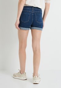 Shorts de mezclilla con un lavado azul oscuro, con los dobladillos enrollados y un diseño de bolsillo trasero estándar. Combinados con zapatillas de color beige claro con acentos dorados.