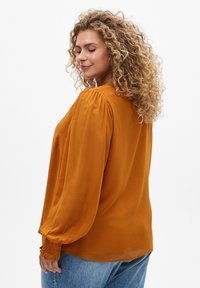 Senfgelbe Bluse aus leichtem Stoff, mit puffigen Ärmeln, raffinierten Manschetten und lockerer Passform, kombiniert mit blauen Jeans.