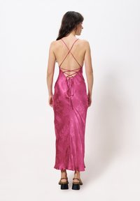 Lang, rugloos jurk van satijn, met een crisscross bandontwerp en vetersluiting aan de achterkant. De kleur is levendig magenta met een subtiele glans.