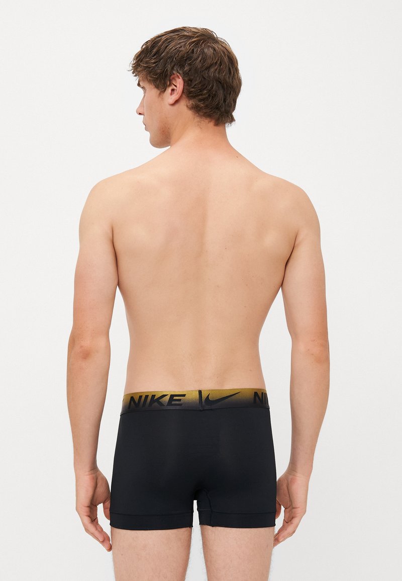 Młody mężczyzna widziany z tyłu, ubrany w czarne bokserki Nike z złotą i czarną gumką w pasie, na jednolitym białym tle.