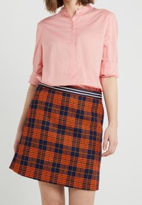 En kvinna i en uppvikt ärm rosa blus instoppad i en hög midja orange och marinblå rutig kjol med ett randigt midjeband.