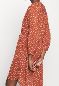 Vestido color óxido con patrón floral blanco, mangas largas, cintura fruncida y ajuste relajado. Tela suave con una textura sutil.