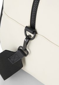 Kermavalkoinen nahkalaukku mustalla vetoketjulla, jossa on metallinen vetoketjun vedin ja musta kangasnauha, joka on kiinnitetty pyöreään metallirenkaaseen.
