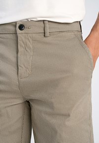Pantalones beige con estampado, hechos de un tejido texturizado, que cuentan con un cierre de botón, bolsillos laterales y un diseño ajustado con pequeños perforaciones.