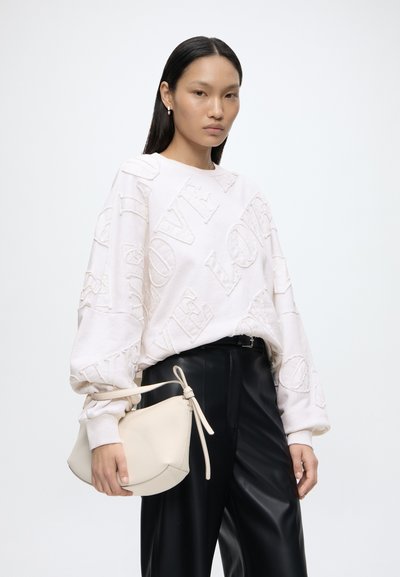Femme aux longs cheveux foncés portant un sweat-shirt blanc texturé avec le texte «LOVE», pantalon noir et tenant un sac à main crème.