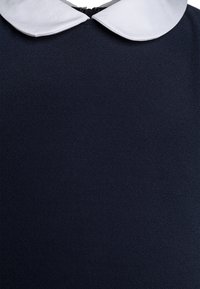 Laivastonsininen mekko, jossa on teksturoitu kangas; siinä on valkoinen pyöreä kaulus ja takana vetoketjusuljenta. Painotus värissä ja suunnittelun yksityiskohdissa.