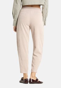 Vrouw in beige ribbroeken en bordeauxrode lakleren platte schoenen met gespen, staand met de rug naar voren gericht op een witte achtergrond.