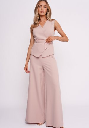 Kobieta ubrana w bezrękawnikowy, jasnoróżowy zawiązywany top oraz pasujące szerokie spodnie, stojąca na jasnym, jednolitym tle.