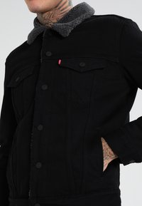 Giacca di jeans nera con colletto in pile grigio, chiusura con bottoni e tasche sul petto. Presenta un design elegante e un logo discreto.