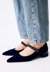 Bailarinas de terciopelo azul marino con punta afilada y un corte redondeado. La correa de tobillo con cuentas negras añade un detalle. Combinadas con unos jeans claros azul celeste remangados.