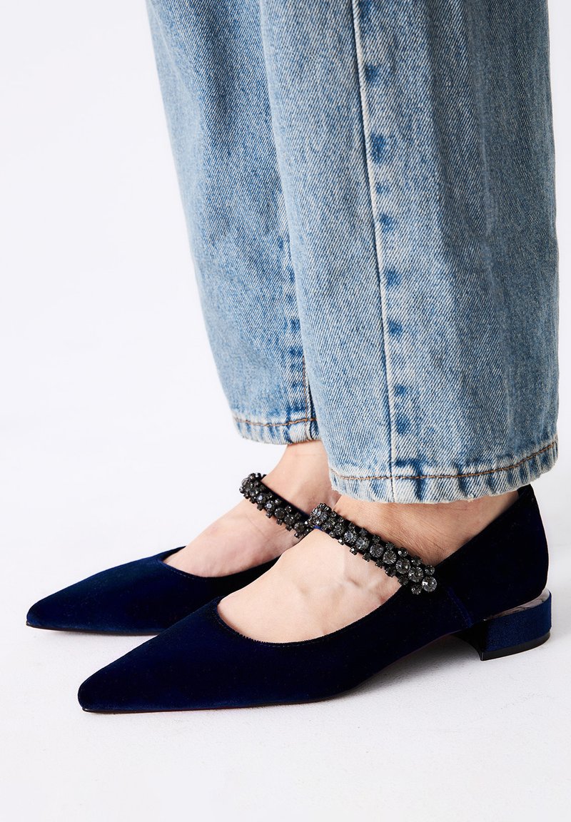 Bailarinas de terciopelo azul marino con punta afilada y un corte redondeado. La correa de tobillo con cuentas negras añade un detalle. Combinadas con unos jeans claros azul celeste remangados.