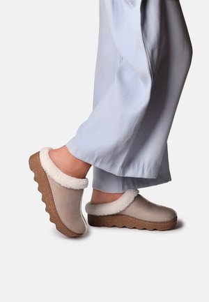 Pieds portant des chaussons beige à enfiler avec une bordure en fourrure blanche et des semelles ondulées marron épaisses, associés à un pantalon ample bleu clair.