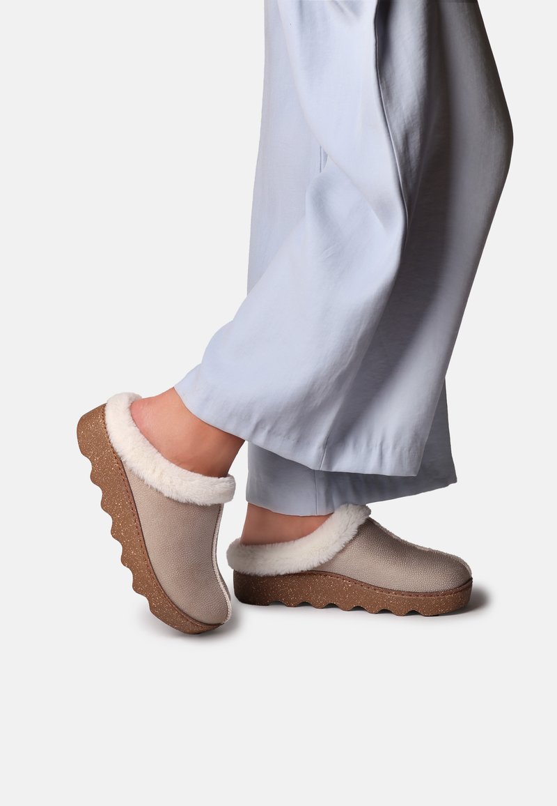 Pieds portant des chaussons beige à enfiler avec une bordure en fourrure blanche et des semelles ondulées marron épaisses, associés à un pantalon ample bleu clair.