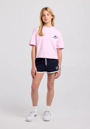 Jonge vrouw staat, draagt een los-roze t-shirt met bloemenprint, marineblauwe shorts met "89", witte sokken en witte Nike sneakers, neutrale achtergrond.