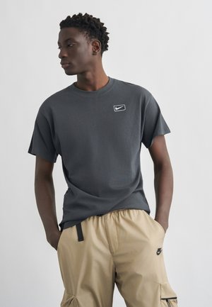 Egy férfi sötétszürke Nike pólóban és bézs Nike cargo nadrágban, zsebre tett kézzel áll, balra néz egy sima háttér előtt.