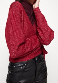 Roter Strickpullover mit V-Ausschnitt und strukturiertem Muster, versehen mit Ballonärmeln. Kombiniert mit schwarzer Lederhose und dezenten metallischen Akzenten.