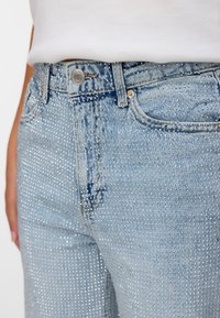 Vaqueros de mezclilla azul claro con un diseño ajustado, adornados con tachuelas plateadas por toda la superficie y un cierre clásico de botón en la cintura.