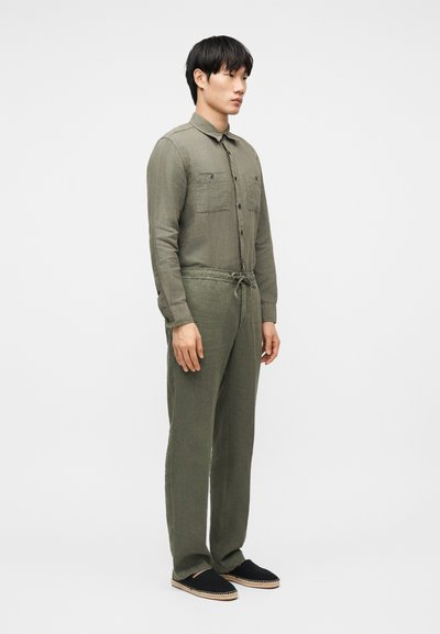 Άνδρας που στέκεται φορώντας πουκάμισο με κουμπιά σε ελιά πράσινο χρώμα και παντελόνι με ελαστική μέση στο ίδιο χρώμα, μαζί με μαύρα παπούτσια χωρίς κορδόνια, μπροστά σε απλό φόντο.