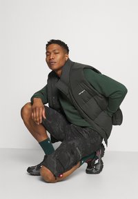 Gilet gonfiato nero sopra un maglione verde, abbinato a pantaloni corti camouflage e sneakers nere. Presenta materiali testurizzati e tasche cargo.