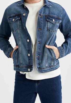 Person trägt eine offene blaue Jeansjacke über einem weißen Hemd, die Hände in den Jackentaschen und kombiniert mit dunkelblauer Jeans.