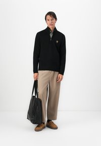 Homem em suéter preto de meia zip, calças largas bege, sapatos castanhos, segurando uma grande bolsa de lona preta, de pé contra um fundo branco simples.