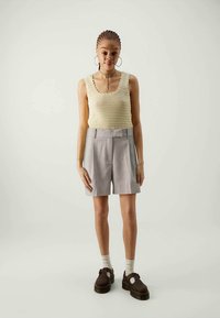 Top de malha em creme com bordas recortadas, combinado com calções cinza claro de corte perfeito. Sapatos castanhos robustos e meias brancas completam o look.