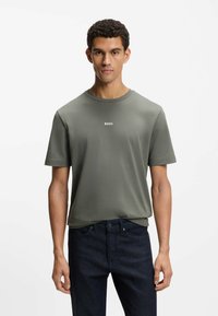 Zielona koszulka z krótkim rękawem i okrągłym dekoltem oraz małym białym logo "BOSS" na piersi, zestawiona z ciemnoniebieskimi dżinsami.