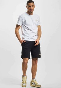 Hvit bomulls t-skjorte med en liten logo, kombinert med svarte shorts. Modellen har på seg høy-top sneakers med et blanding av gull- og hvite detaljer.