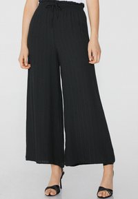 Pantalones de pierna ancha negros con una tela texturizada, cintura elástica con cordón y diseño en capas. Combinados con tacones negros con tiras.