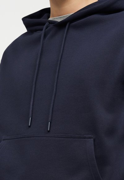 Άτομο που φοράει μπλε-σκούφο φούτερ με κορδόνια πάνω από γκρι πουκάμισο, εμφανίζοντας το πάνω μέρος του κορμού και λεπτομέρειες της μπροστινής τσέπης.