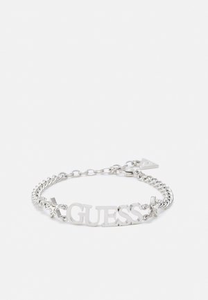 Silbernes Kettenarmband mit "GUESS" Buchstaben und Sternanhängern auf weißem Hintergrund, mit einem kleinen dreieckigen Logo-Verschluss.