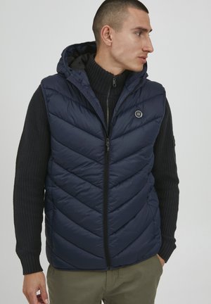 Navy blue puffer vest with chevron quilted pattern, zippered front, and hood. Worn over a black ribbed sweater, features a logo accent.