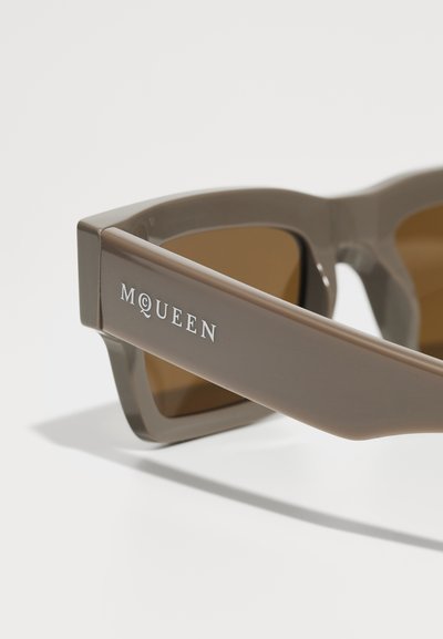 Occhiali da sole in plastica marrone con lenti rettangolari, caratterizzati dal nome del marchio "MQUEEN" in bianco sulla stanghetta. Finitura texturizzata e stanghette larghe.
