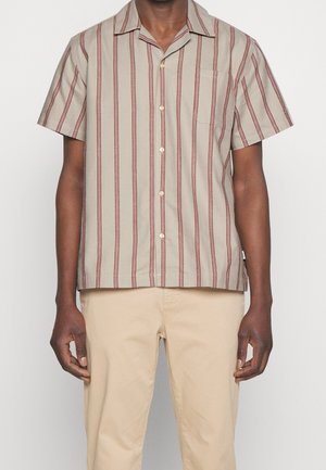 Mężczyzna w beżowej koszuli z krótkim rękawem zapinanej na guziki z pionowymi czerwonymi paskami oraz jasnobeżowych spodniach, stojący na jednolitym tle.