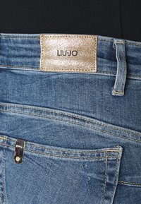 Vaqueros de mezclilla en un lavado azul claro con una etiqueta de logo dorado brillante. Cuenta con cinco bolsillos, costuras visibles y un corte recto clásico.