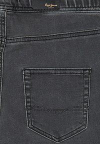 Los jeans de mezclilla negros presentan un bolsillo trasero distintivo con un detalle de costura. La tela tiene una textura suave con un ligero brillo.