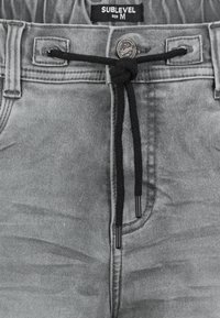 Gray denim pants with an elastic waistband and black drawstring. Features a round logo tag and stitched details along the pockets. Size M.