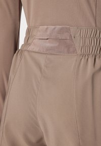 Pantaloni sportivi marroni chiari con una cintura elastica testurizzata e un pannello in rete a contrasto. Tessuto liscio con cuciture dettagliate.