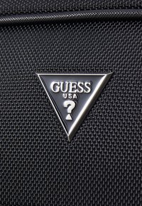 Tessuto nero con texture, dotato di un distintivo metallico triangolare, che presenta "GUESS USA" e un design con il punto interrogativo in bianco al centro.