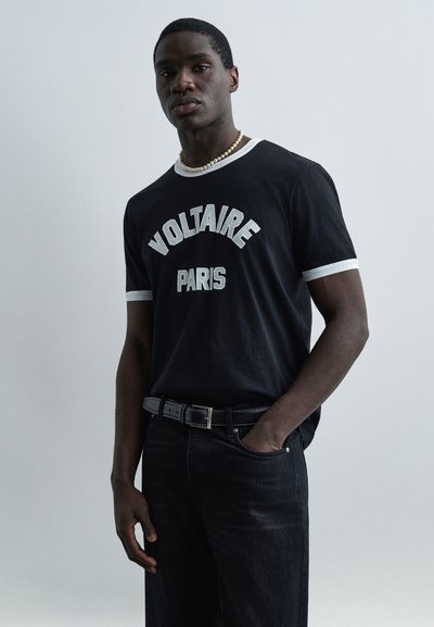 Άνδρας με μαύρο T-shirt "Voltaire Paris" με λευκή επένδυση, μαύρα τζιν, μαύρη ζώνη και κολιέ με μαργαριτάρια, στέκεται μπροστά σε απλό φόντο.