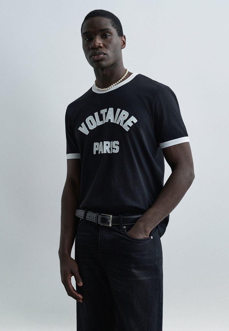 Άνδρας με μαύρο T-shirt "Voltaire Paris" με λευκή επέν δυση, μαύρα τζιν, μαύρη ζώνη και κολιέ με μαργαριτάρια, στέκεται μπροστά σε απλό φόντο.