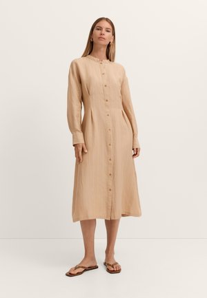 Frau steht und trägt ein beigefarbenes, langärmeliges Midikleid mit Knopfleiste, vertikalen Streifen und braunen flachen Sandalen.