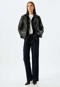 Giacca in pelle nera lucida con colletto ripiegato, abbinata a pantaloni in denim scuri a vita alta e tacchi a punta neri.