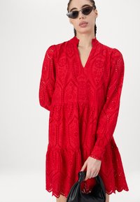 Röd, broderad klänning med ett lager-på-lager design, med V-ringning och långa puffärmar. Bärs med svarta accessoarer.