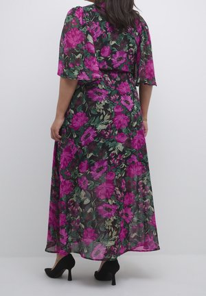 Kobieta w długiej czarnej sukience z jaskrawym fioletowym kwiatowym wzorem i falbanowymi rękawami, w połączeniu z czarnymi butami na wysokim obcasie.