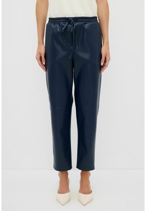 Woman wearing navy blue faux leather pants with drawstring waist and white pointed-toe heels, standing against a plain background.