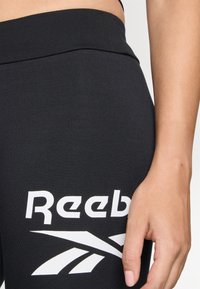 Svarte treningsleggings med bred midje, med hvit "Reebok" logo og aksendesign på nedre del av benet, glatt tekstur.