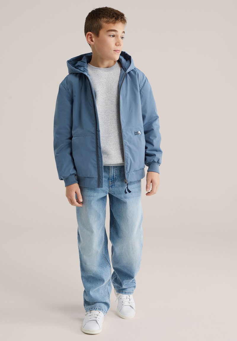 Modrá lehká bunda s kapucí, zapínáním na zip a elastickými manžetami, nošená přes šedé tričko. Kombinováno s lehkými džíny a bílými teniskami.