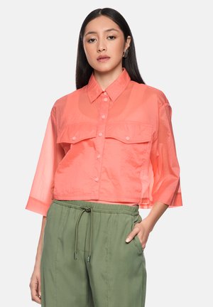 Woman with long dark hair wearing a coral buttoned shirt with chest pockets and olive green drawstring pants, hand in pocket.